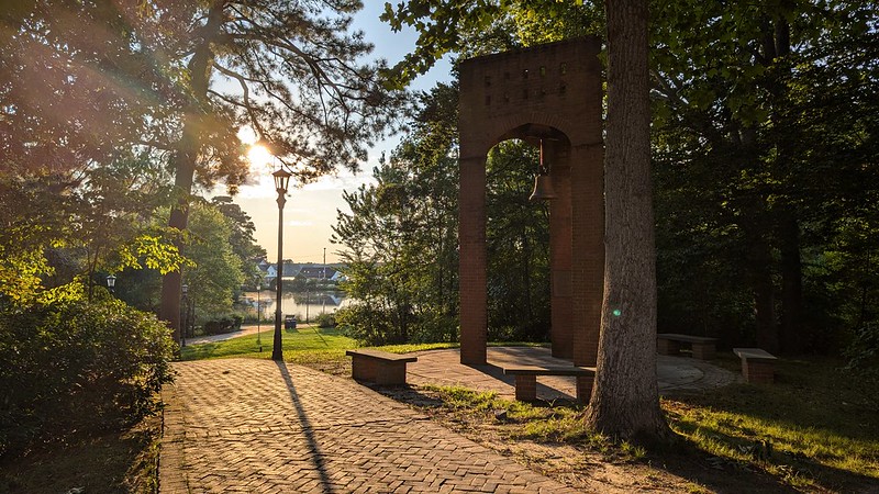 Brick arch with a bell stands in a shaded park area near a paved walkway, trees, benches, and a lamppost, with a river and sunlight in the background.