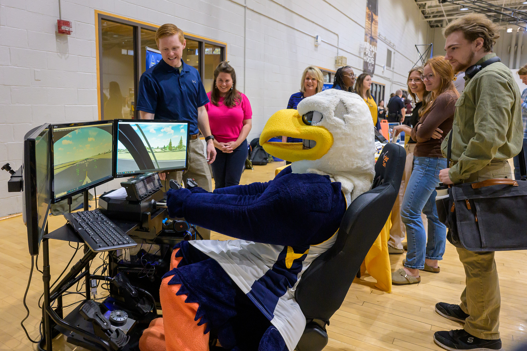 A person in an eagle mascot costume sits at a racing simulator setup, while several people stand nearby watching in a gymnasium setting.