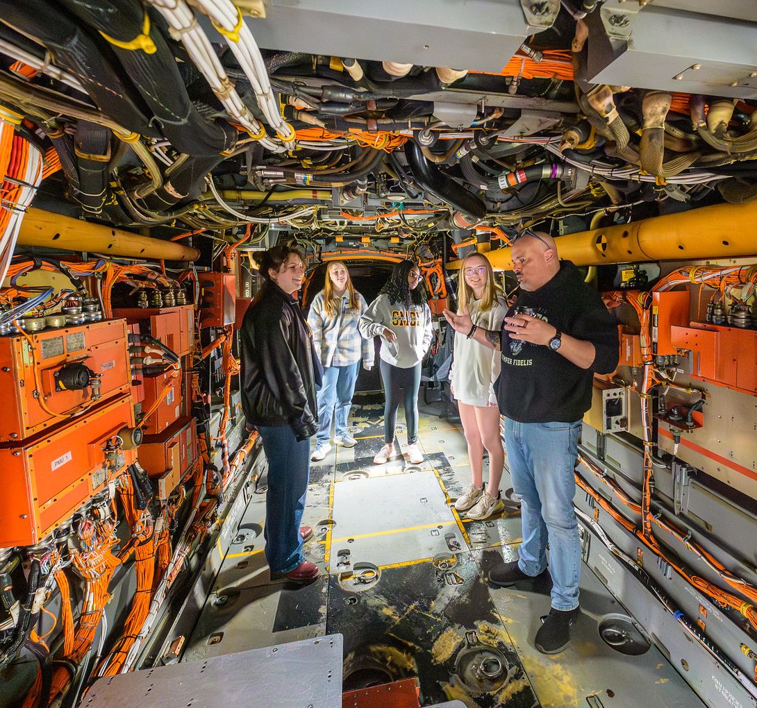 A group of people stands inside a narrow, exposed mechanical area of an aircraft, surrounded by wires, pipes, and orange equipment panels.
