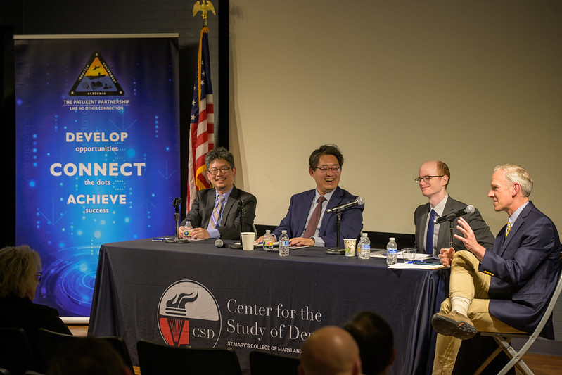Four men in suits sit at a panel table with microphones, speaking to an audience. A banner and an American flag are in the background. The table has a "Center for the Study of Democracy" sign.