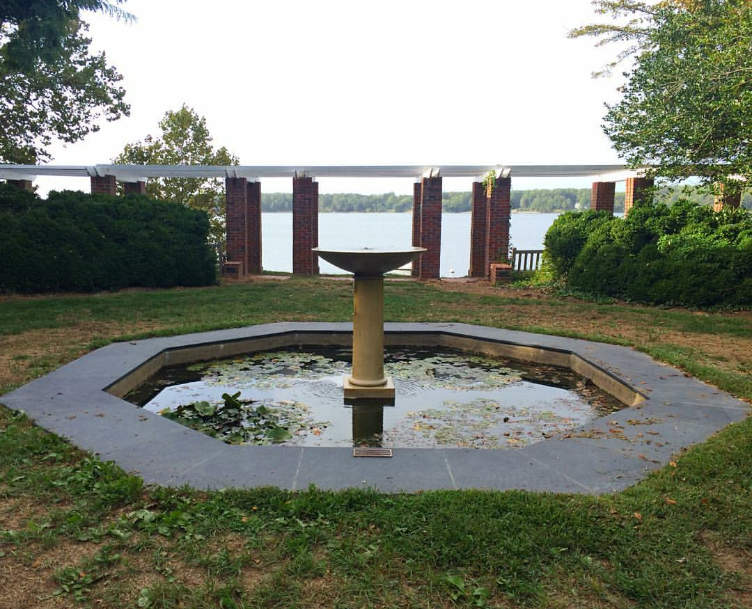 Octagonal pond with a central pedestal birdbath, surrounded by grass and hedges—this tranquil haven is framed by brick columns, with a lake in the background, creating a setting that whispers of garden wonders.