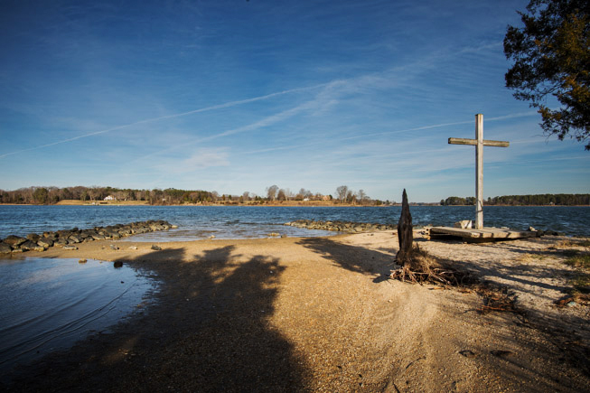 A large wooden cross stands as one of nature’s wonders on a sandy lakeshore under a blue sky, with water, rocks, and trees in the background.