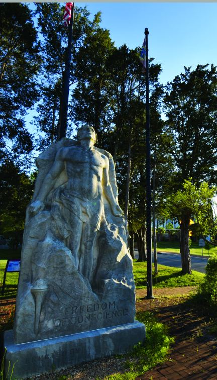 Stone statue of a standing figure with the words “FREEDOM OF CONSCIENCE” engraved; flanked by two flagpoles and surrounded by trees in an outdoor setting, this remarkable monument stands as one of the quiet wonders nestled within nature’s embrace.