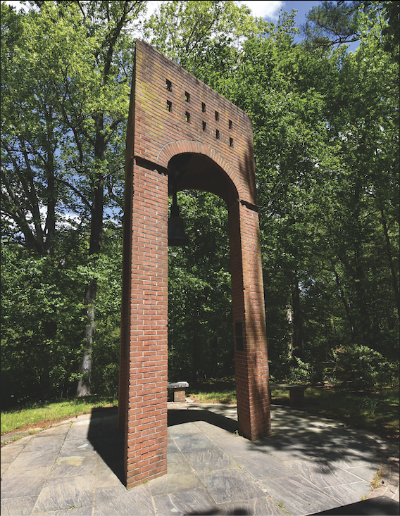 A tall brick arch, one of the local wonders, with a hanging bell stands on a stone platform surrounded by trees and greenery under a partly cloudy sky.