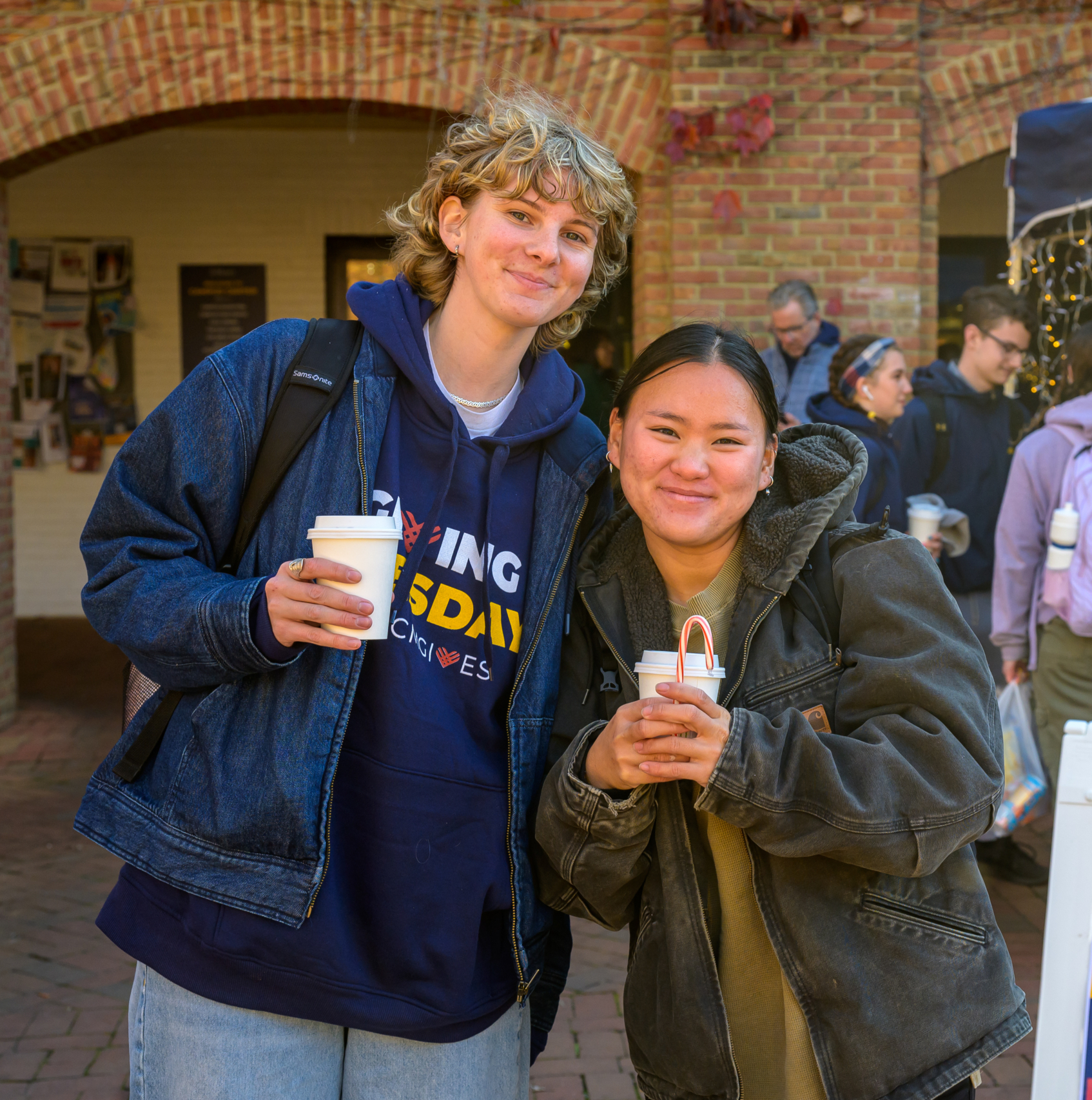 Two people stand smiling while holding coffee cups at an outdoor event. They are dressed warmly in jackets. A sign nearby reads "BeCounted at the Allan Challenge Voting Station." People and a brick building are in the background.