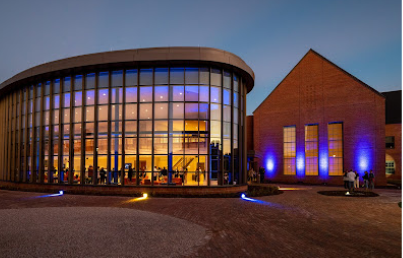 A modern glass-fronted building and a brick building are illuminated with blue lights at dusk, with people visible inside and outside.