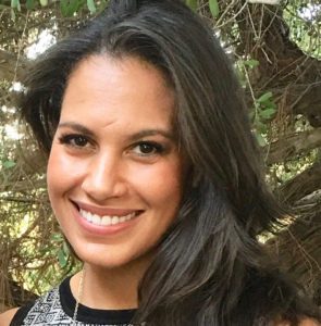 A woman with long dark hair smiles in front of trees and greenery.