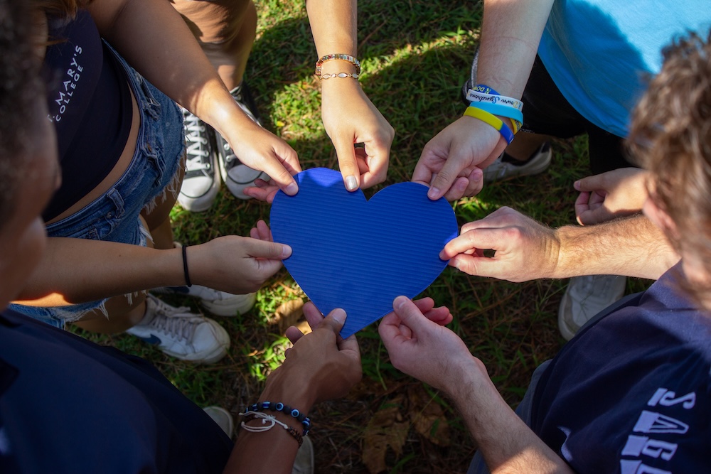 A group of people standing in a circle outside, each holding a piece of a blue heart-shaped cutout in the center.