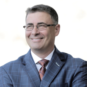 A man wearing glasses, a blue plaid suit, and a striped tie is smiling while looking to the side against a white background.