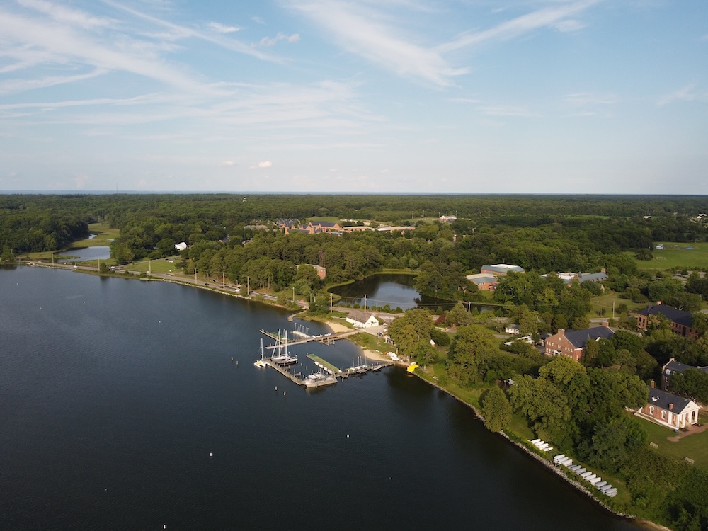 Aerial view of a waterfront marina with sailboats docked, surrounded by green trees, buildings, and open landscape under a partly cloudy sky.