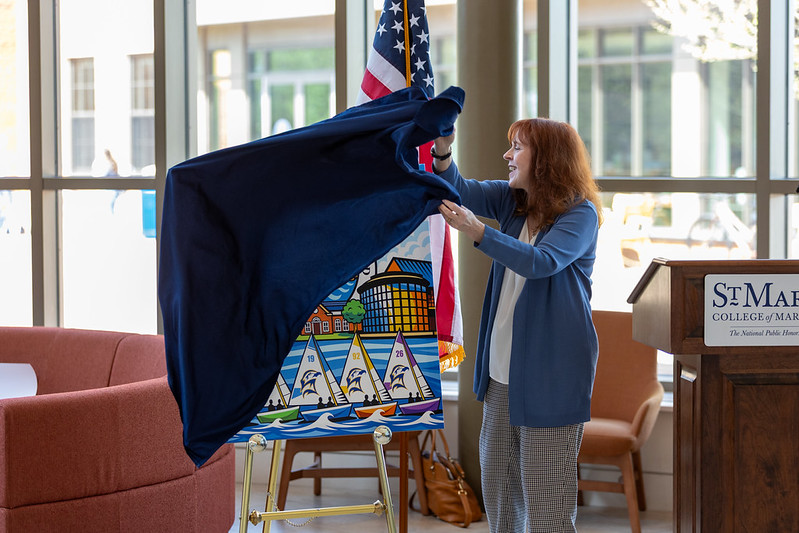 A woman unveils a colorful painting featuring sailboats and buildings at an indoor event near a podium labeled "St. Mary's College of Maryland.
