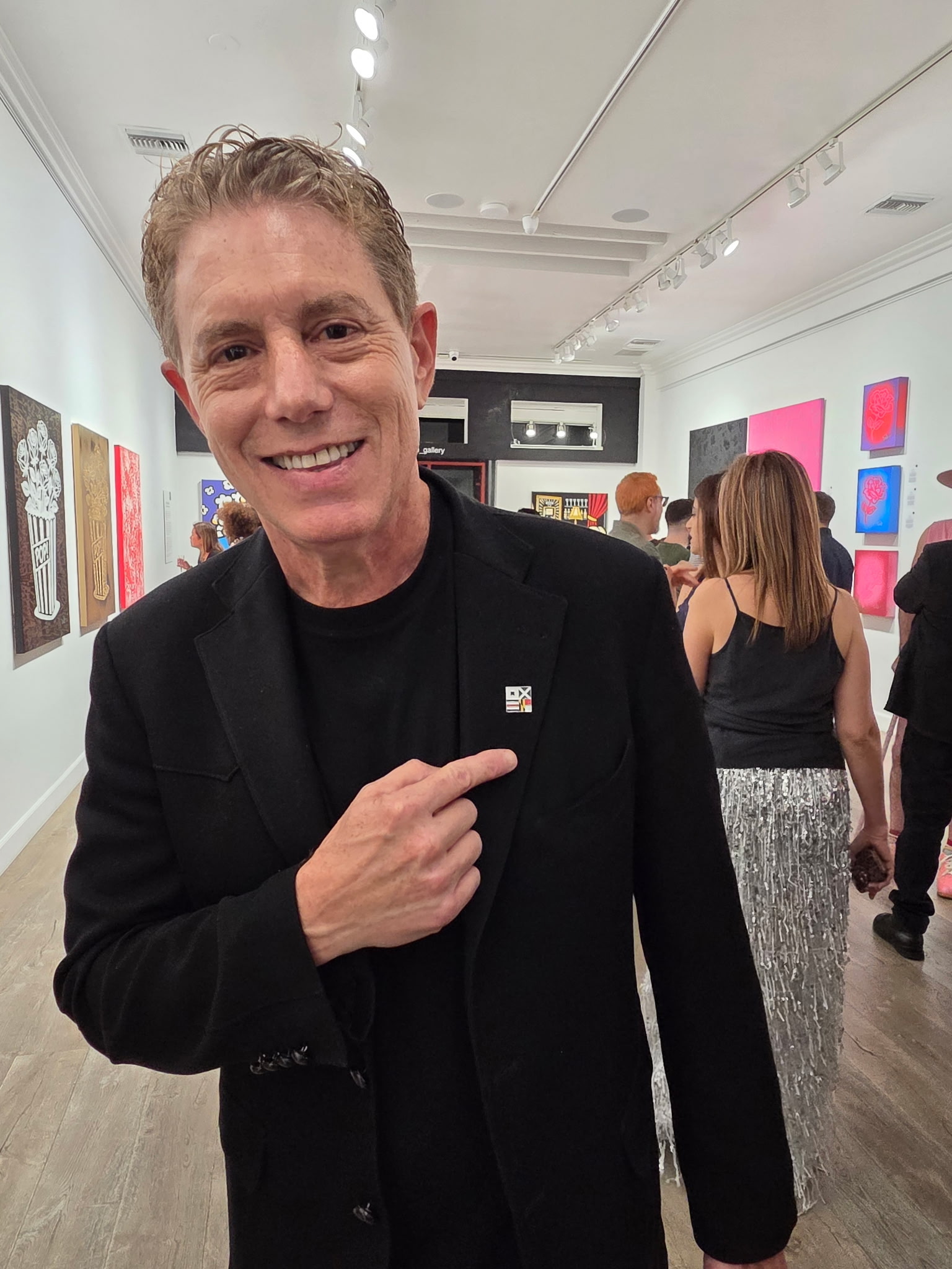A man in a black suit points to a pin on his lapel while standing in an art gallery with several people and colorful artworks in the background.