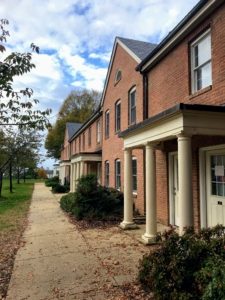 A row of brick townhouse buildings with white columns line a sidewalk. There are green shrubs in front of the townhouses, and trees in the background under a partly cloudy sky. Fallen leaves are scattered on the ground.