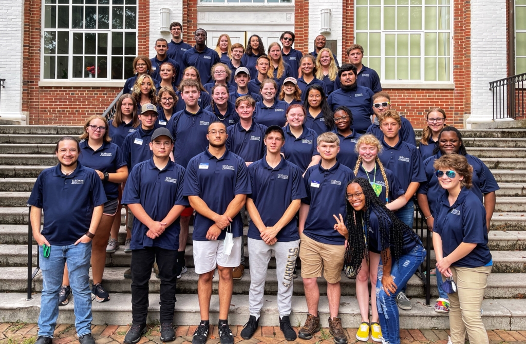 A group of people, wearing navy blue shirts, stand together on outdoor steps in front of a brick building with large windows. Some are smiling and posing with peace signs.