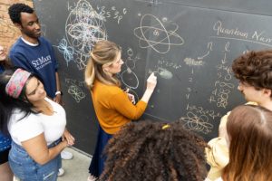A teacher in an orange sweater draws complex quantum diagrams on a chalkboard, surrounded by attentive students. The board features mathematical symbols and intricate shapes. They appear engaged in a lively discussion.