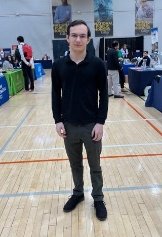 A young man in glasses stands on a gym floor at an indoor event with tables, banners, and people in the background.