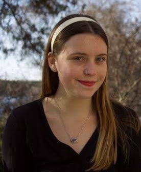 A young woman with long hair, wearing a black top and headband, stands outdoors in front of trees on a sunny day.