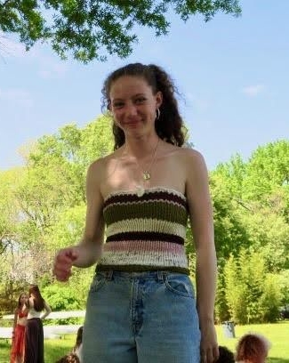 A young woman smiling at the camera. Trees, a green building, and a red-and-white tent are visible in the background.