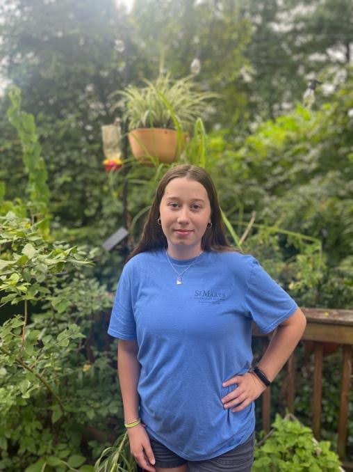 A young woman stands outdoors in front of lush green plants, wearing a blue T-shirt and shorts, with one hand on her hip and a neutral expression.