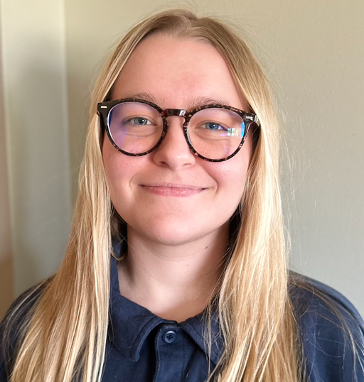 A person with long blond hair and glasses is standing indoors, facing the camera, and smiling slightly. They are wearing a dark blue shirt.