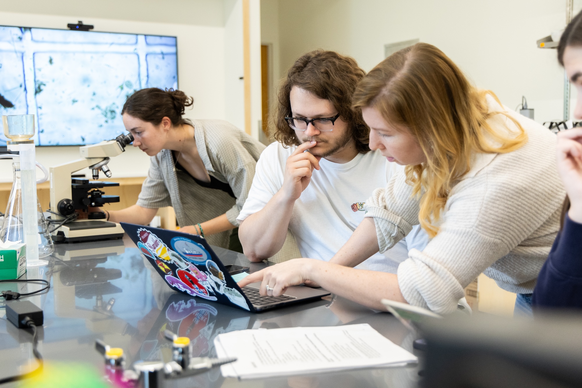 Three people work in a laboratory; one uses a microscope while two others focus on a laptop covered with stickers, with documents and lab equipment on the table.
