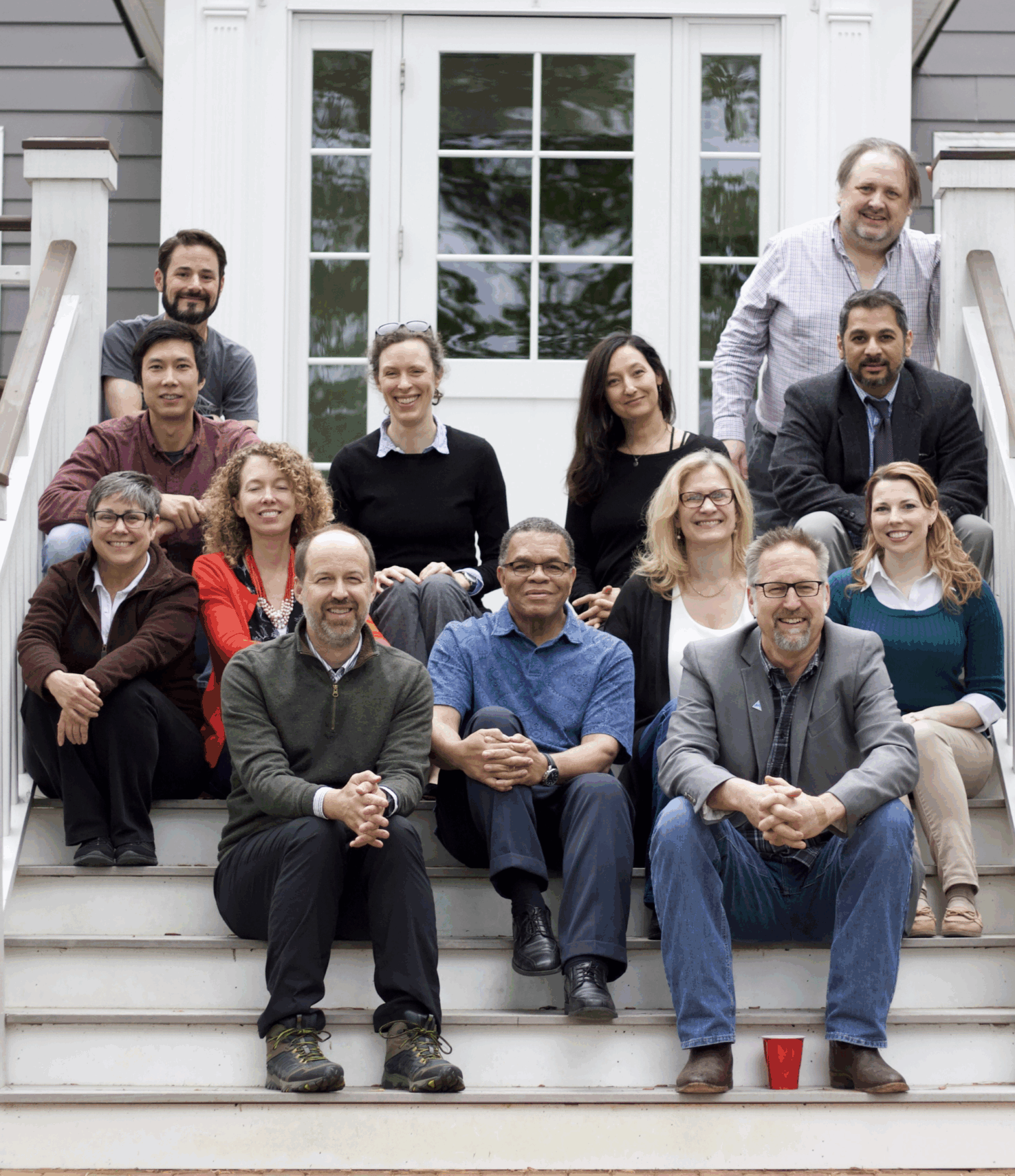 A group of fourteen adults sits and stands on outdoor steps in front of a house, facing the camera and posing for a group photo.