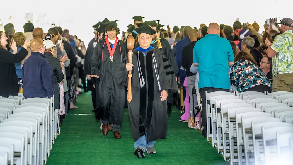A faculty member in academic regalia leads a procession of graduates down an aisle lined with seated and standing attendees at a graduation ceremony.