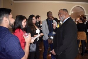A group of people in formal attire are interacting at a networking event. One person is holding a drink and smiling, while others listen and engage in conversation, holding notebooks and wearing name tags. The setting is a warmly lit room.