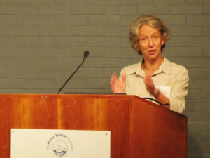 A person with short, light hair, wearing a light-colored shirt, is speaking at a wooden podium with a microphone. The podium has an emblem on it, and there's a gray brick wall in the background.