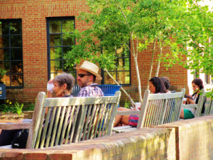 People sitting on wooden benches by a brick wall and building, with greenery around. Some are drinking and talking, including a person in a hat and sunglasses. Sunlight casts warm shadows, creating a relaxed outdoor setting.
