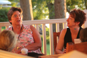 Two women sit on a porch, engaged in conversation. The woman on the left wears a floral sleeveless top and glasses, while the woman on the right wears a black sleeveless top. There are trees and a railing in the background.