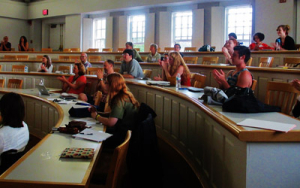 A lecture hall with rows of students sitting at desks. The room is well-lit, with large windows on one side. Some people are clapping, while others are focused on laptops and notebooks.
