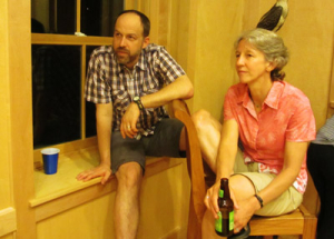 A man and a woman are seated in a room with wooden walls. The man is sitting on a windowsill with one leg on a chair, and the woman is seated with a bottle in hand. Both appear to be attentively watching something.
