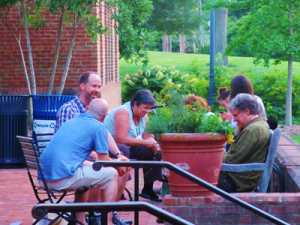 Five people are gathered outside on a brick patio, sitting around a large potted plant. They are engaged in conversation. Greenery and a brick building are in the background.