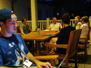 A group of people sit around a wooden table on a porch at night. One person in the foreground, wearing a cap backwards, types on a laptop. Others are engaged in conversation, with drinks on the table. The scene is dimly lit.