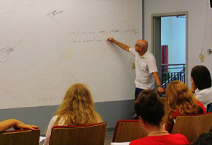 A man is writing equations on a large whiteboard in a classroom setting. Several people are seated, facing the board and taking notes. The room has a relaxed, educational atmosphere.