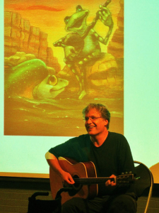 A person sitting and playing a guitar, smiling, in front of a screen displaying an illustration of two frogs. One frog is playing a guitar on a rock, while the other is in the water, set against a sunset background.