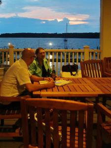 Two people sit at a wooden table on a patio overlooking a lake at dusk. A few sailboats are visible on the water. The sky is a mix of blue and orange hues, with trees lining the horizon. The patio is lit softly by nearby lights.