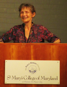An elderly woman stands at a podium labeled "St. Mary's College of Maryland." She is smiling and wearing a purple patterned blouse, speaking into a microphone with a brick wall in the background.