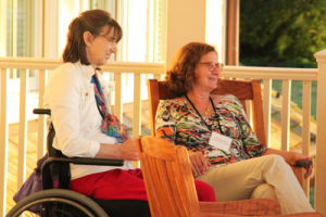 Two women sitting on a porch, one in a wheelchair and the other on a wooden chair. They are smiling and appear to be having a pleasant conversation. The setting has warm lighting and a relaxed atmosphere.