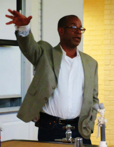 A person in a gray blazer and white shirt gestures while speaking. They are standing near a lectern with a small faucet visible. The background includes a yellow brick wall and a whiteboard.