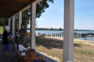 People are gathered on a covered porch with columns, overlooking a beach and a pier extending into a calm lake. It's a sunny day, and some individuals are conversing while others relax in chairs, enjoying the scenic view.
