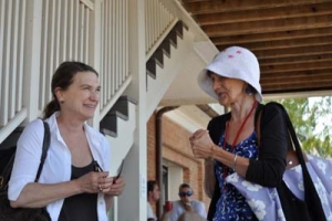 Two women stand and chat under a staircase. One wears a white shirt and holds an object, while the other, in a blue dress with a white sun hat, carries a patterned tote bag. People are blurred in the background.