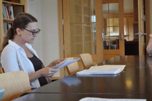 A woman with glasses and a ponytail sits at a conference table, reading documents. She is wearing a black top and white cardigan. The room has a wooden door and bookshelves in the background.