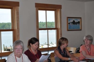 Four women are sitting at a table in a room with large windows. Two of them are writing notes. The windows overlook a lake with docks and trees. A framed painting hangs on the wall.
