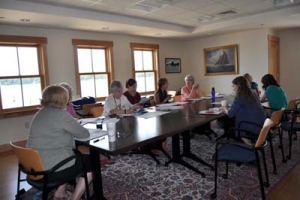 A group of people sitting around a long, rectangular table in a conference room with large windows and framed art on the walls, engaged in discussion with papers and notebooks. The room is well-lit and has a patterned rug.