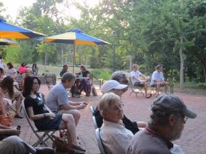 A group of people sit on chairs in an outdoor area with brick flooring. Sunlight filters through trees in the background. Blue and yellow umbrellas provide shade, and some individuals are using their phones. The scene feels relaxed and social.