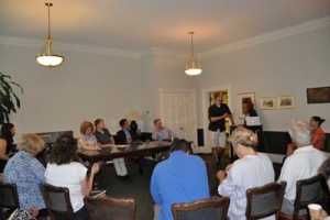 A group of people are seated around a long table in a meeting room. One person stands at the front, speaking and gesturing. The room has light-colored walls and framed pictures. Ceiling lights are on, and a laptop is open on a pedestal.