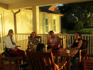 Four people are sitting on a porch around a wooden table, engaging in conversation. The setting is warmly lit by the evening sun. A coffee cup and bag are on the table. In the background, a grassy area and buildings are visible.