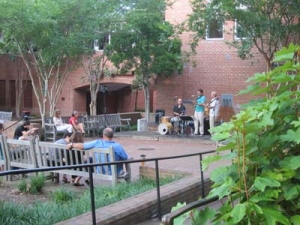 A small jazz band performs in a courtyard surrounded by trees and brick buildings. People sit on benches, listening to the music. The scene is relaxed and inviting, with greenery adding to the ambiance.